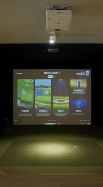 Indoor golf simulator setup with a projected screen displaying options for practice, courses, games, tournaments, and online play.