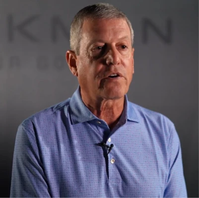 Middle-aged man in a light blue polo shirt speaking on camera with a lapel microphone against a dark blurred backdrop.