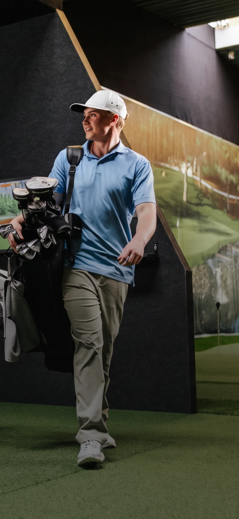 A man in a blue polo and white cap carries a golf bag, walking on indoor turf with a golf simulator background.