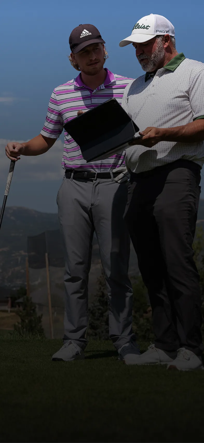 Two men on a golf course reviewing a tablet. One holds a golf club, and they stand against a backdrop of mountains and a clear sky.