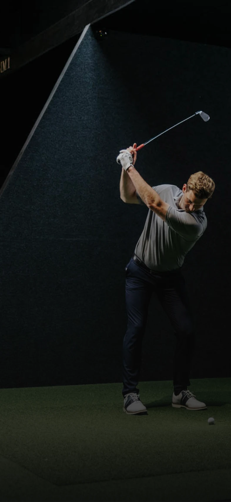 Golfer in a gray shirt and dark pants swings a club indoors, poised to hit a golf ball on green turf under focused lighting.