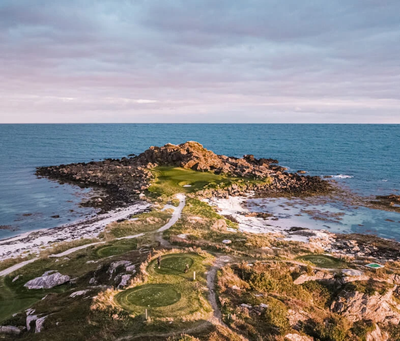 Aerial view of a rocky coastline with a narrow path leading to a grassy area, surrounded by the ocean under a cloudy sky.