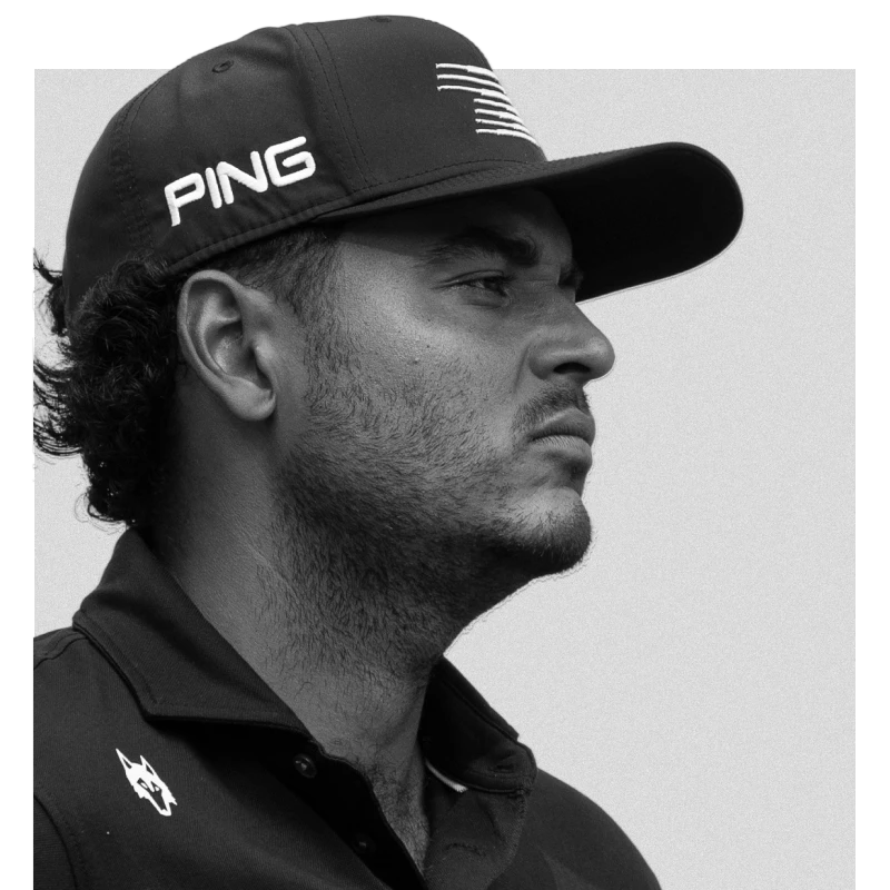 Man in a black cap with "PING" logo, wearing a dark collared shirt, looking serious and gazing to the side; black and white photo.
